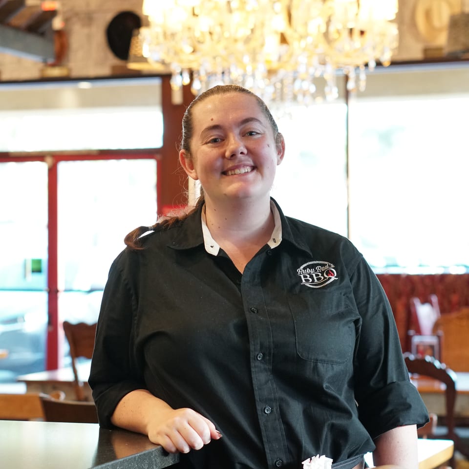 Smiling Ruby Red’s BBQ server in a black uniform stands inside the warmly lit restaurant beneath a crystal chandelier.